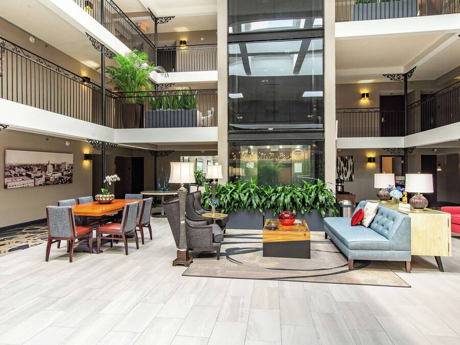 Un hall d’entrée spacieux avec un plafond haut, un coin repas avec une table et des chaises, un canapé et une table basse. Des plantes et une grande fenêtre se trouvent au centre.