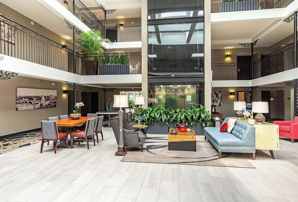 A spacious lobby with a high ceiling, a dining area with a table and chairs, a sofa, and a coffee table. There are plants and a large window in the center. - The Burgundy Hotel, Tapestry Collection by Hilton (Little Rock)