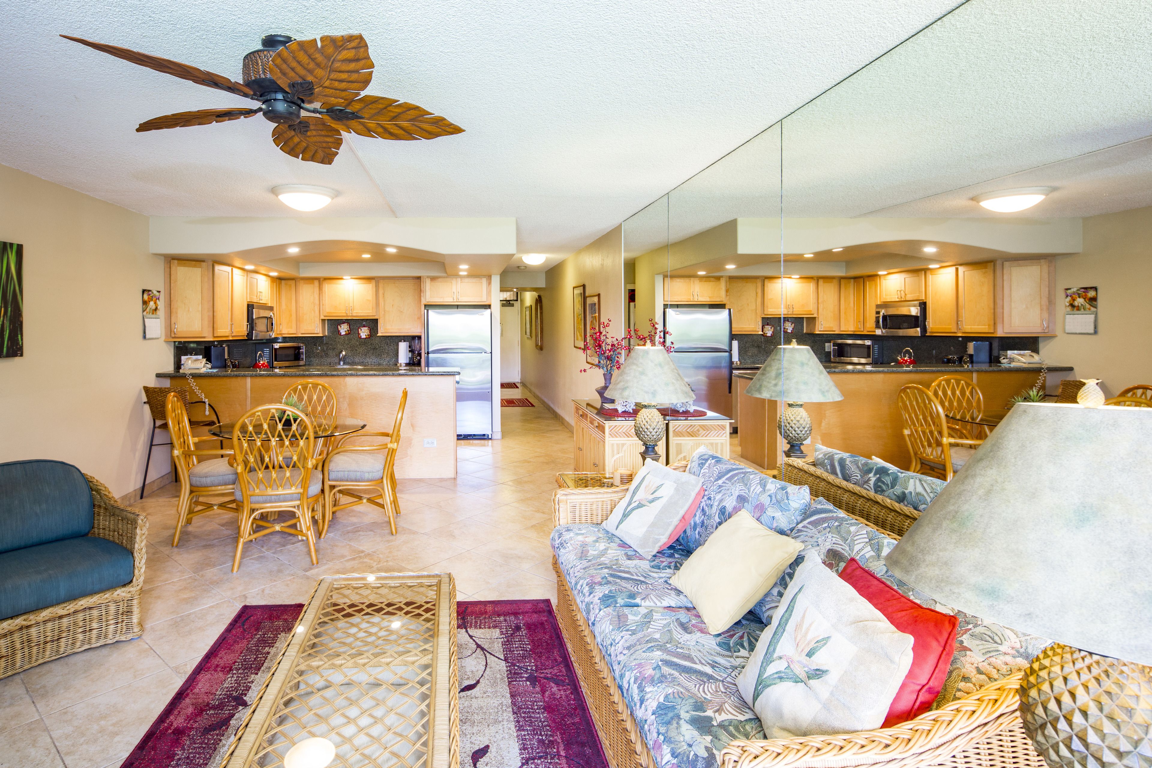 A spacious living room with a large sofa, a dining area with chairs, a kitchen with wooden cabinets, and a ceiling fan.