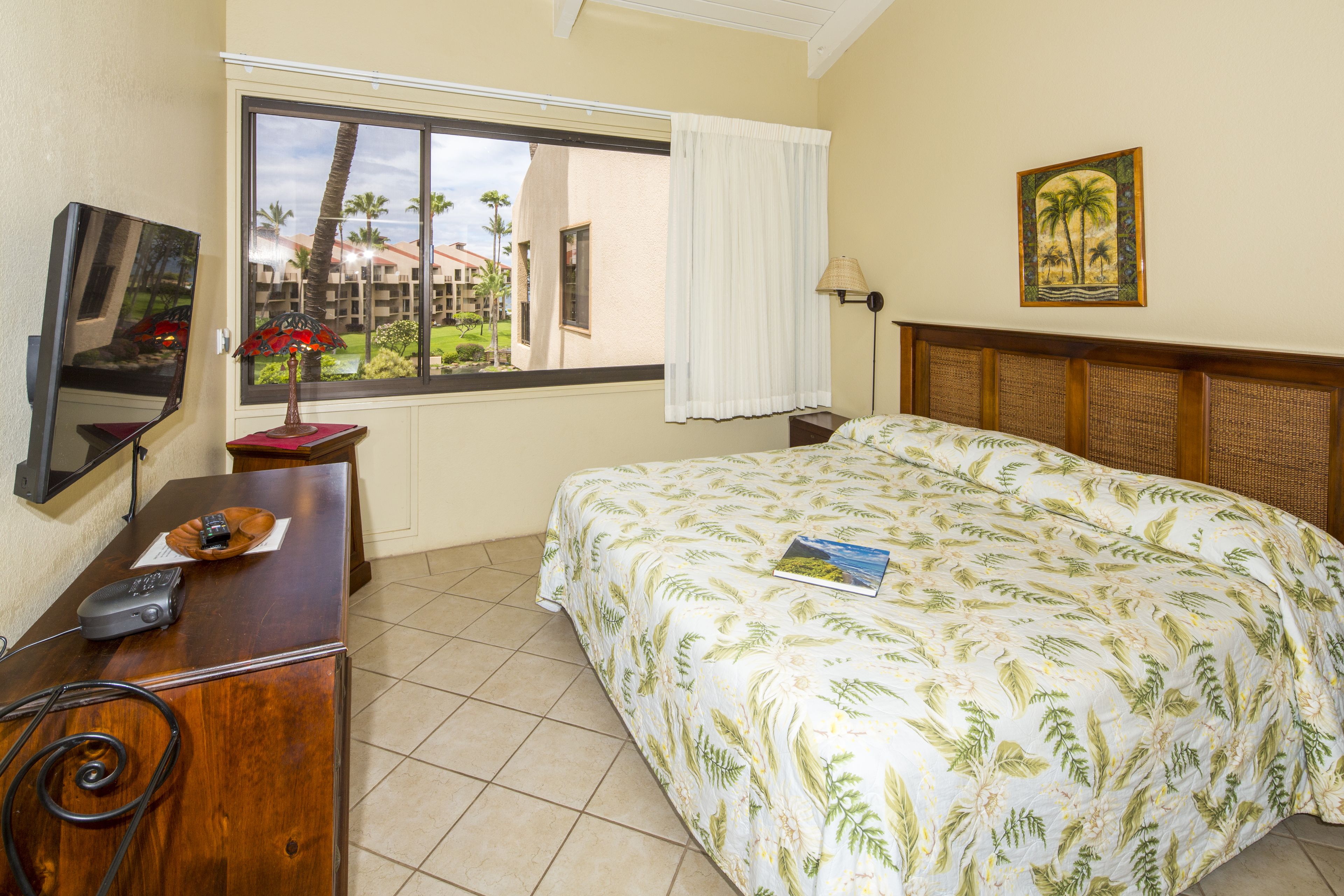 A hotel room with a bed, a desk with a lamp, a television, and a window with a view of palm trees and buildings.