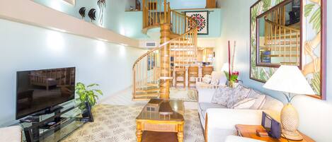 Un salon spacieux doté d’un grand téléviseur à écran plat, d’une table basse en verre et d’un canapé confortable. Un escalier en bois mène à un niveau supérieur où se trouve un balcon.