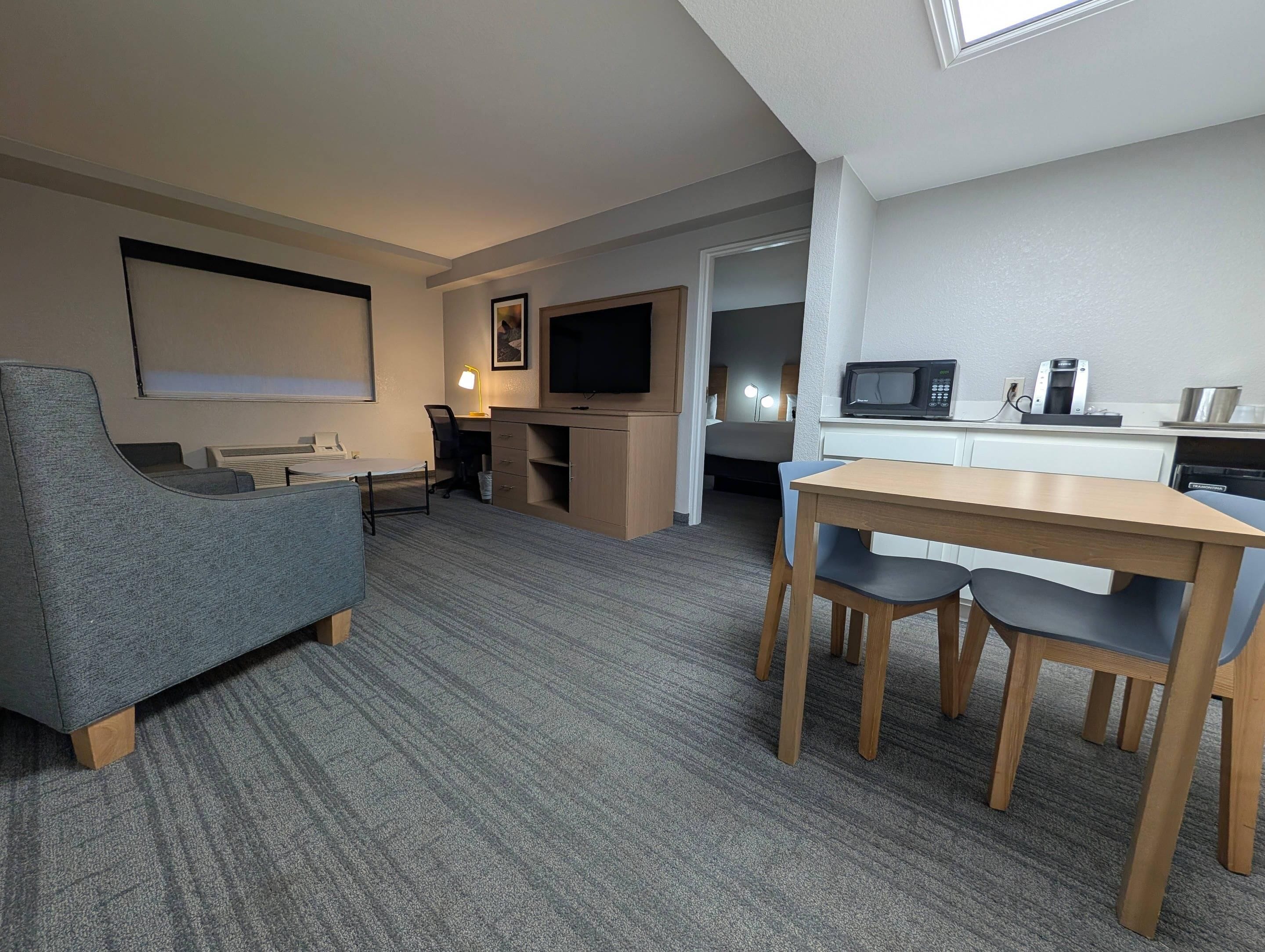 A hotel room with a gray sofa, a wooden dining table with chairs, a kitchenette, a TV, and a large window.