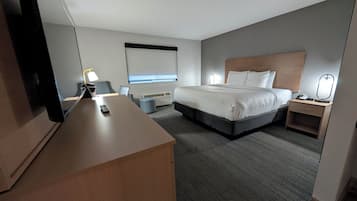 A hotel room with a large bed, a desk, a chair, a bedside table with a lamp, a small trash bin, and a wall-mounted TV.