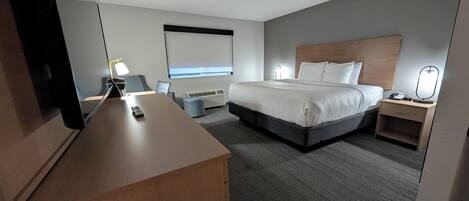 A hotel room with a large bed, a desk, a chair, a bedside table with a lamp, a small trash bin, and a wall-mounted TV.