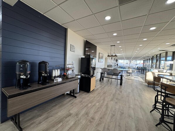 A modern coffee station with multiple coffee makers, a seating area with tables and chairs, and a large window providing a view of the outside.