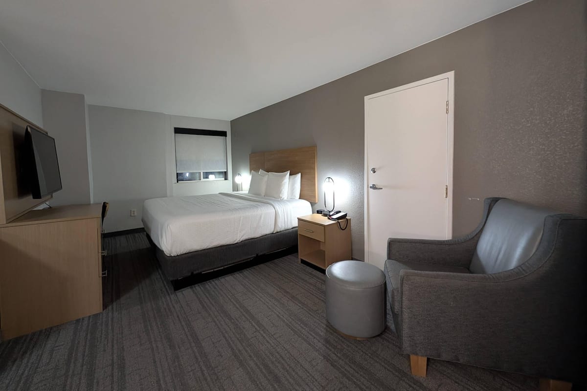 A hotel room with a bed, bedside table, lamp, TV, and a grey armchair.