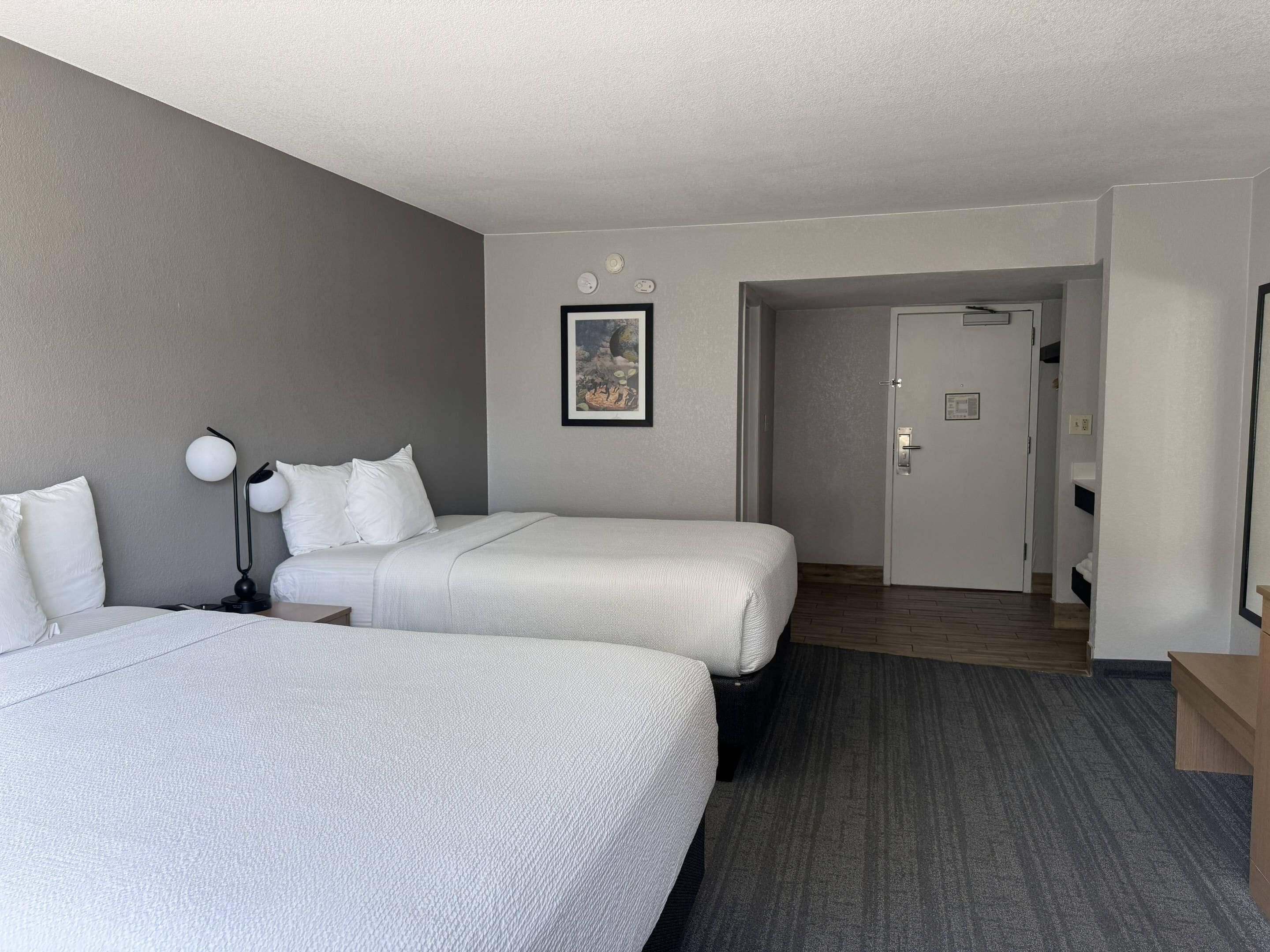 A hotel room with two beds, a nightstand, a lamp, a framed picture on the wall, and a door leading to another room.