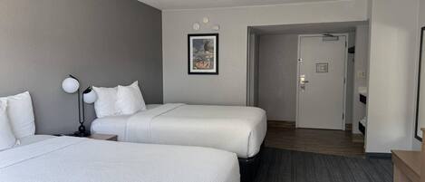 A hotel room with two beds, a nightstand, a lamp, a framed picture on the wall, and a door leading to another room.