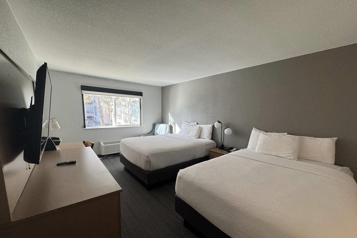 A hotel room with two beds, a desk, a television, and a window with a view of buildings.