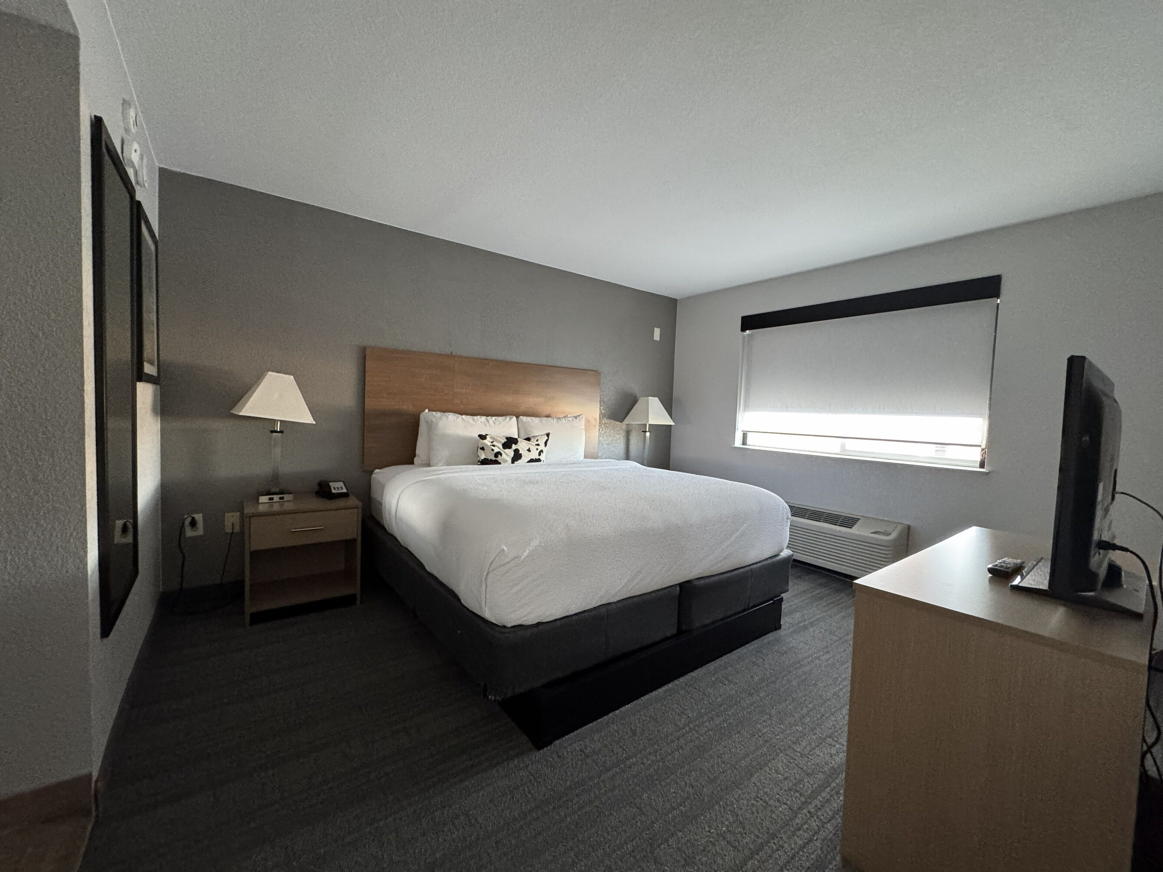 A hotel room with a large bed, bedside tables with lamps, a desk with a television, and a window with blinds.