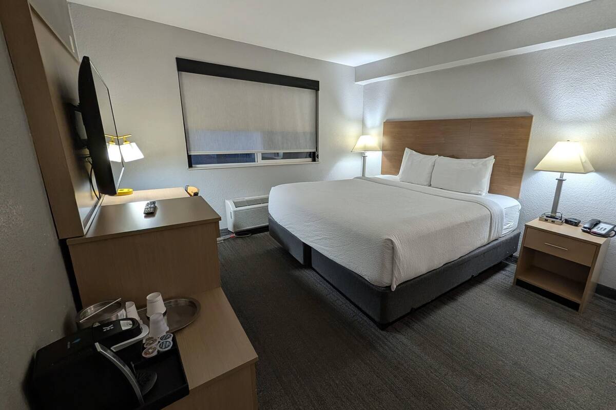 A hotel room with a large bed, bedside tables with lamps, a flat-screen TV, a mirror, and a window with blinds.