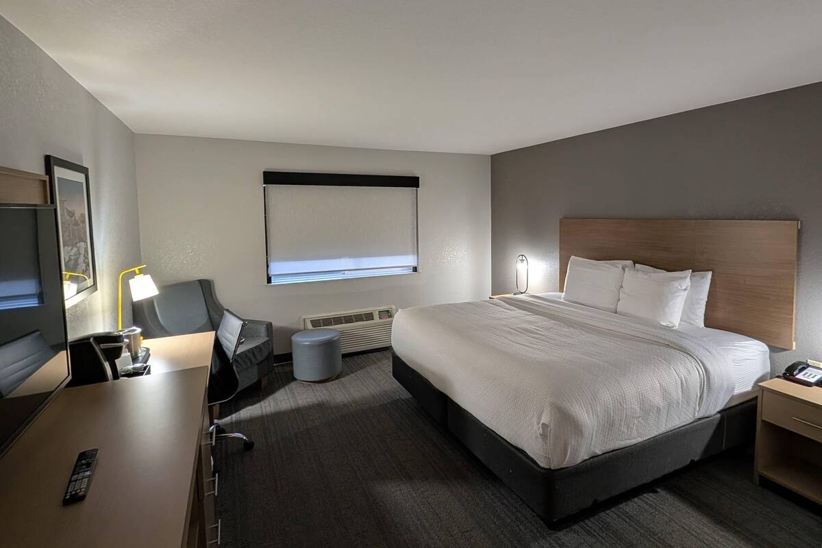 A hotel room with a bed, bedside table, lamp, TV, and a window with blinds.