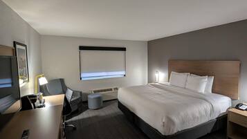 A hotel room with a bed, bedside table, lamp, TV, and a window with blinds.