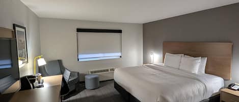 Quarto de hotel com cama, mesa de cabeceira, luminária, televisão e janela com persianas.