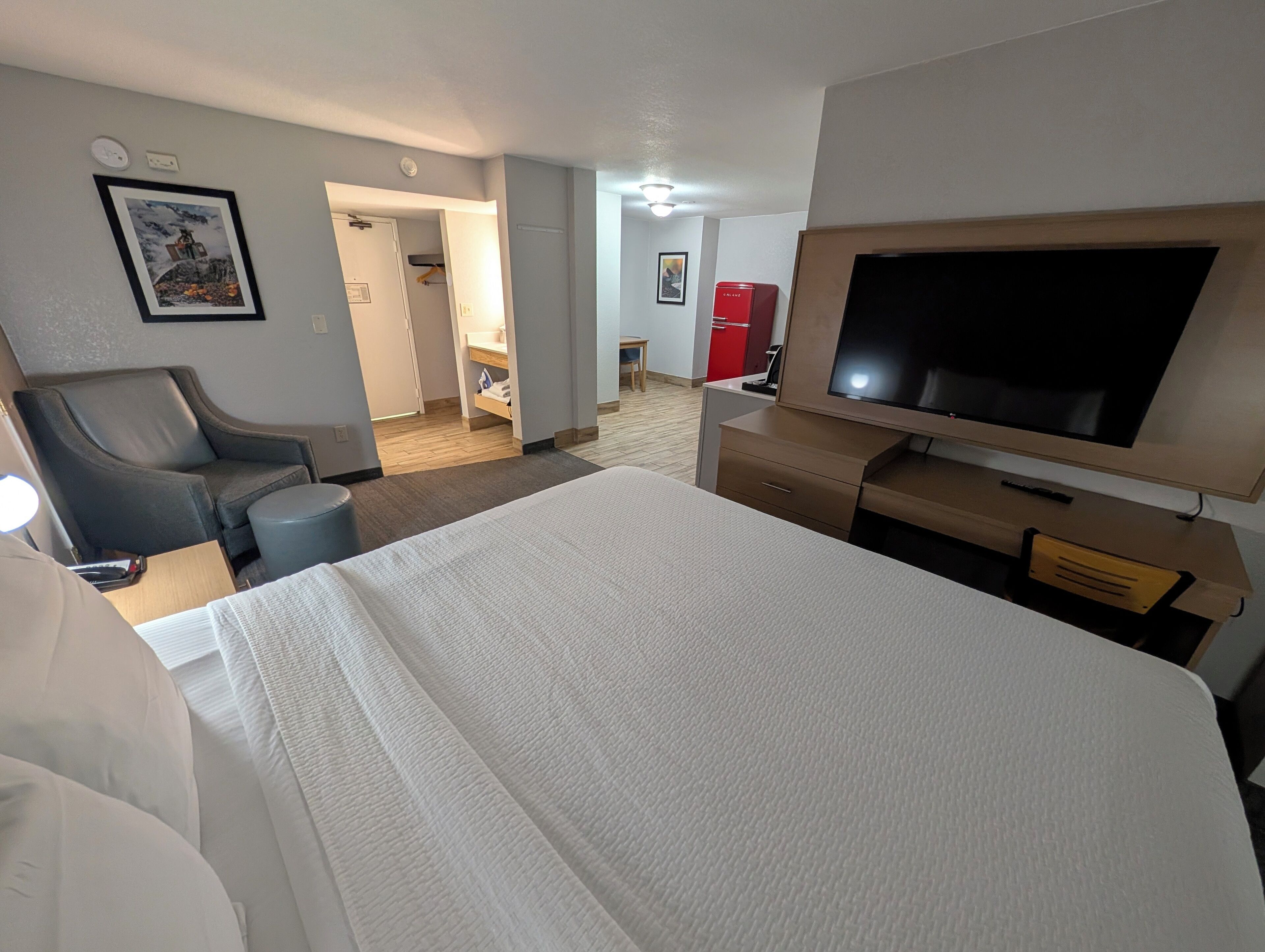 A hotel room with a large bed, a television, a desk, and a chair. There is a door leading to a bathroom and a kitchenette.