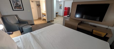 Une chambre d’hôtel avec un grand lit, une télévision, un bureau et une chaise. Une porte donne accès à une salle de bain et à une kitchenette.