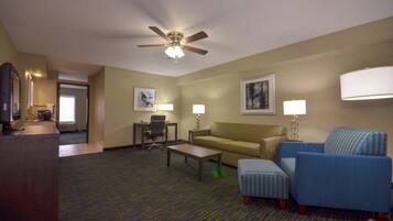 A hotel room with a ceiling fan, a sofa, a coffee table, a desk with a chair, a television, and artwork on the walls.