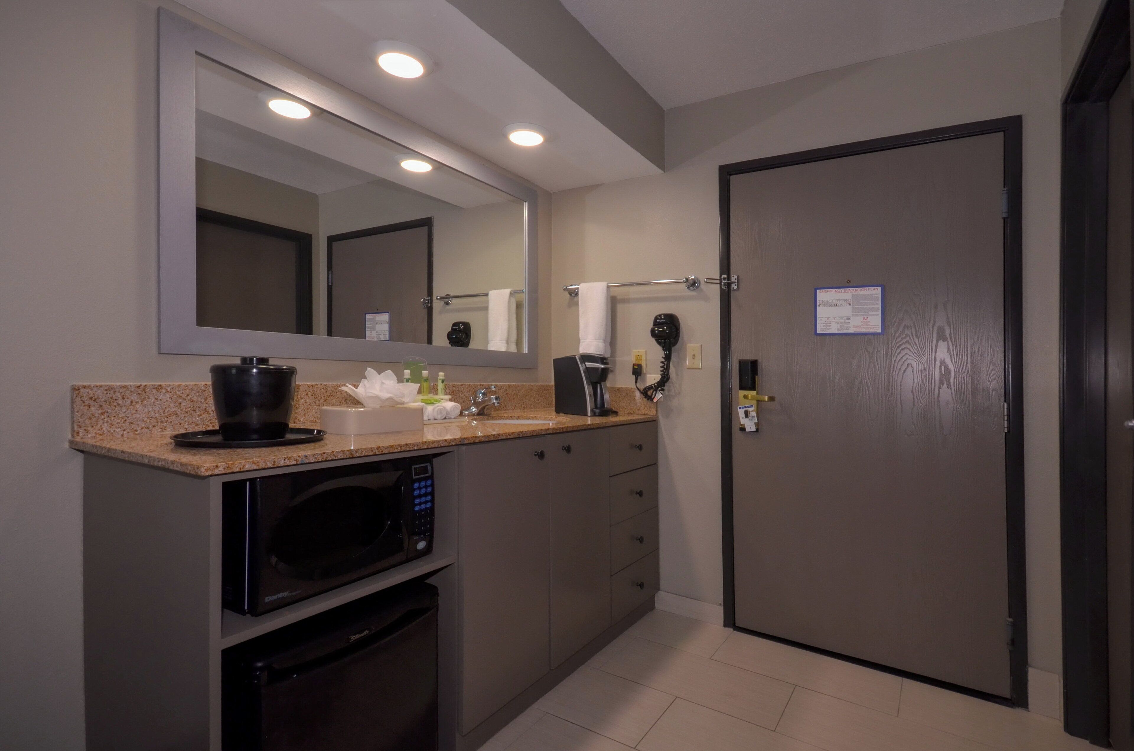 A compact hotel bathroom with a sink, mirror, towel rack, hairdryer, and a door with an emergency evacuation plan sign.
