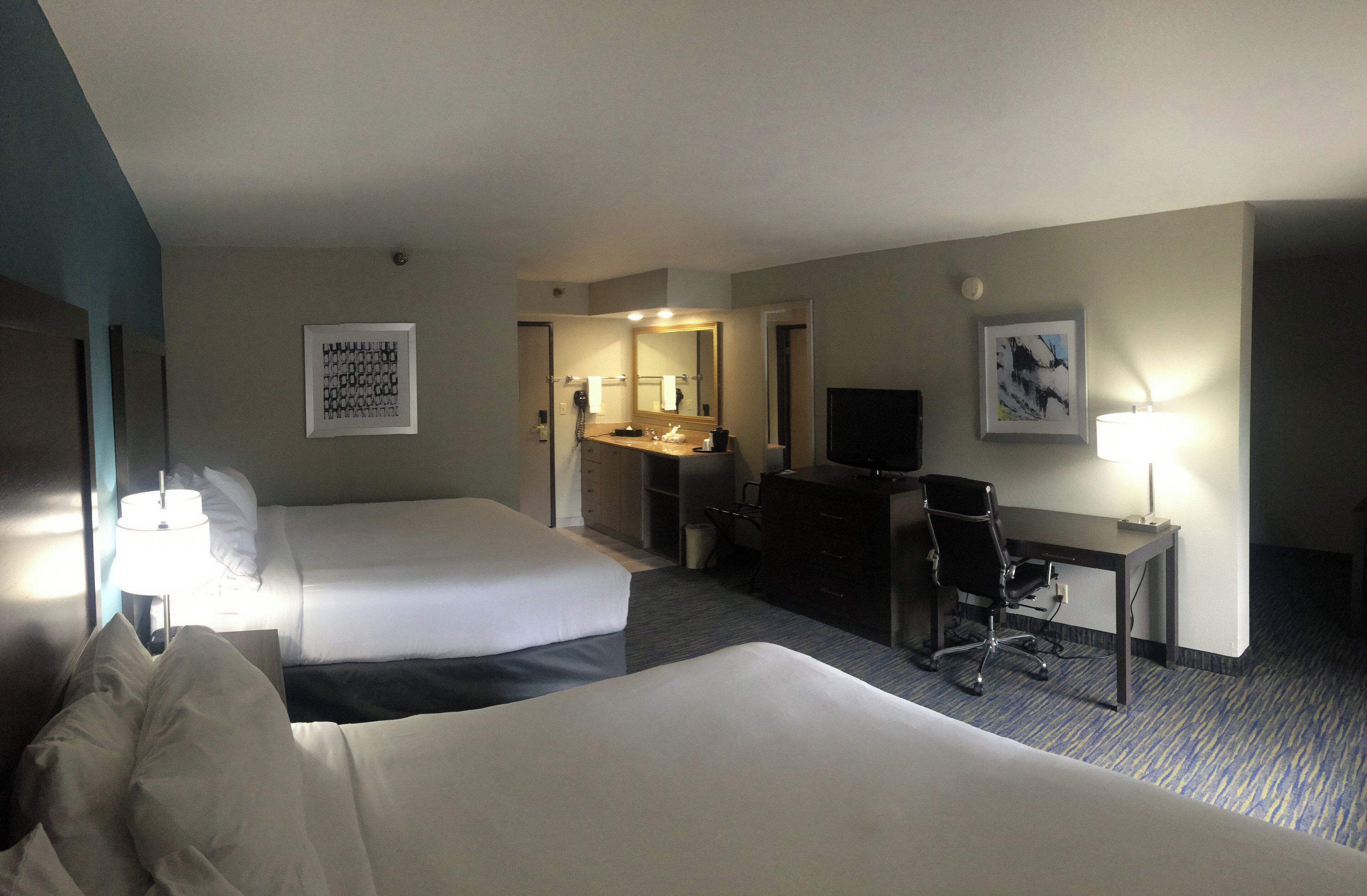 Habitación de hotel con dos camas, un escritorio con silla, televisor y baño con espejo y lavabo.
