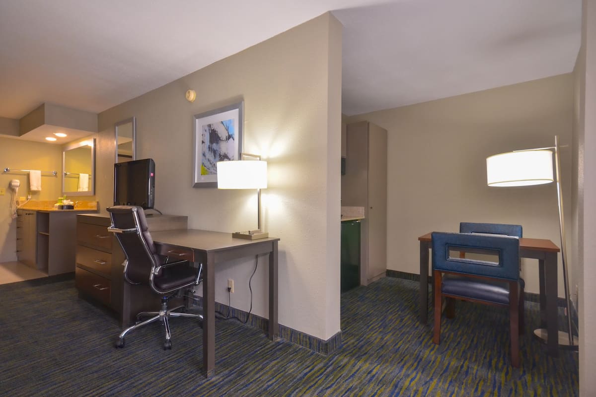 A hotel room with a desk, two chairs, a lamp, a mirror, a television, and a carpeted floor.
