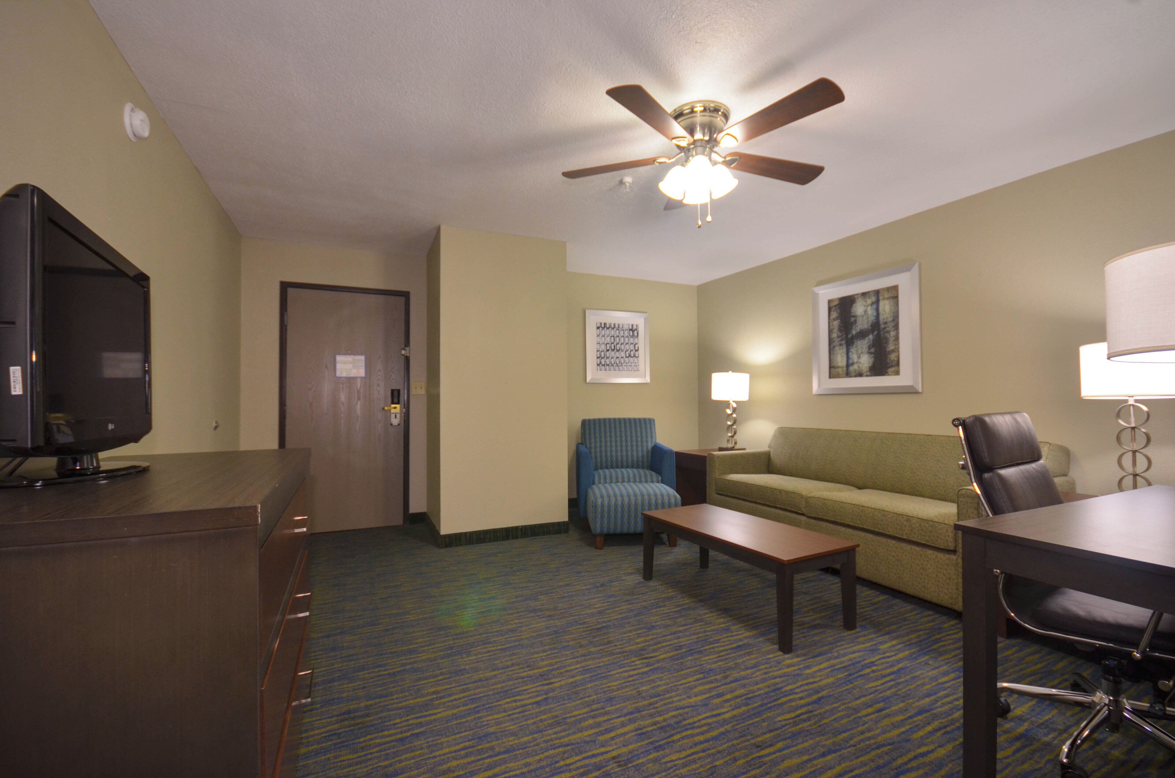 A hotel room with a television, a ceiling fan, a sofa, a coffee table, and a desk with a chair.