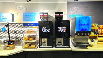 A coffee shop counter with multiple coffee machines, a bakery display, and a sign that reads "MUST HAVE COFFEE".