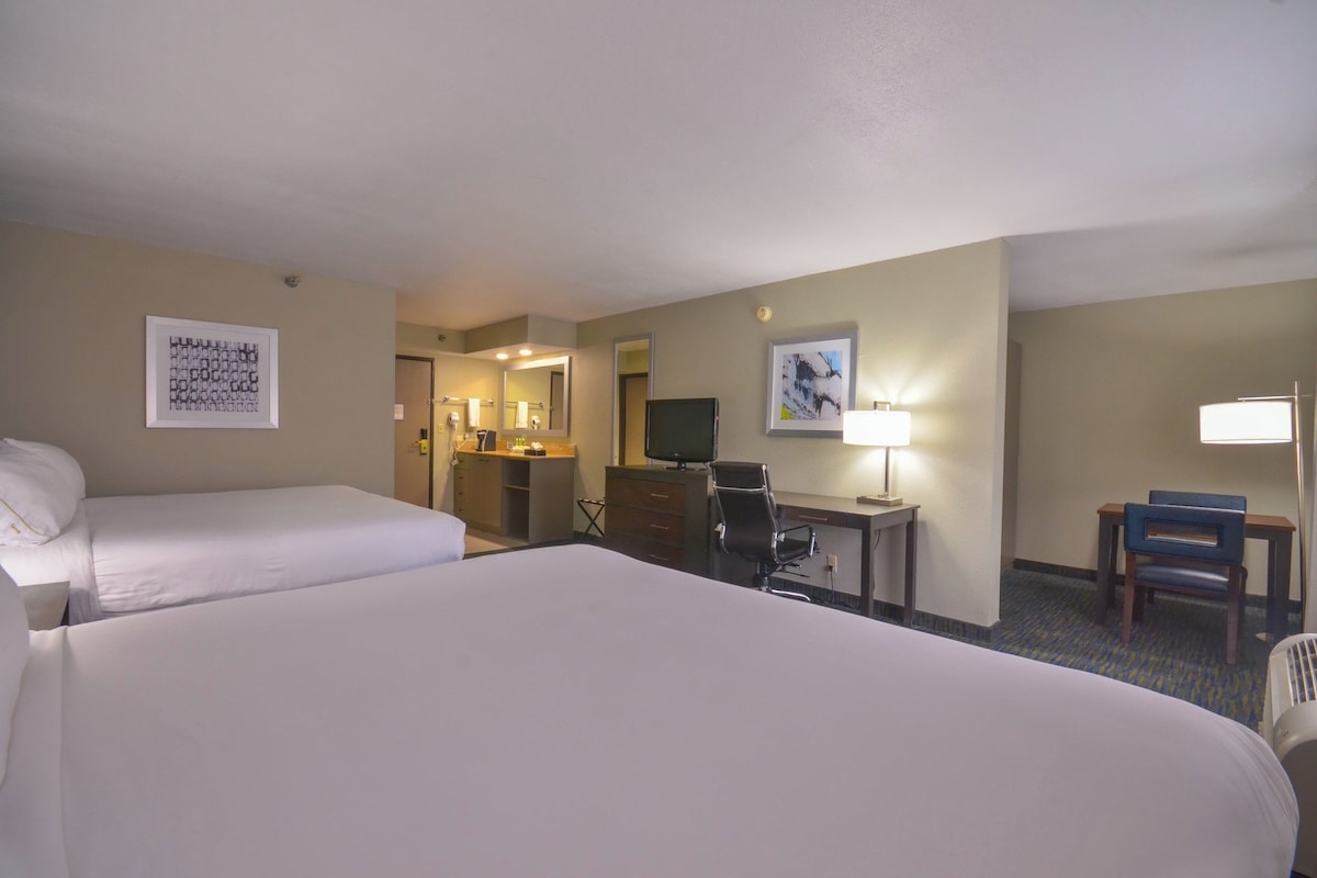 Quarto de hotel com duas camas, uma escrivaninha com cadeira, televisão e banheiro visível por uma porta aberta.