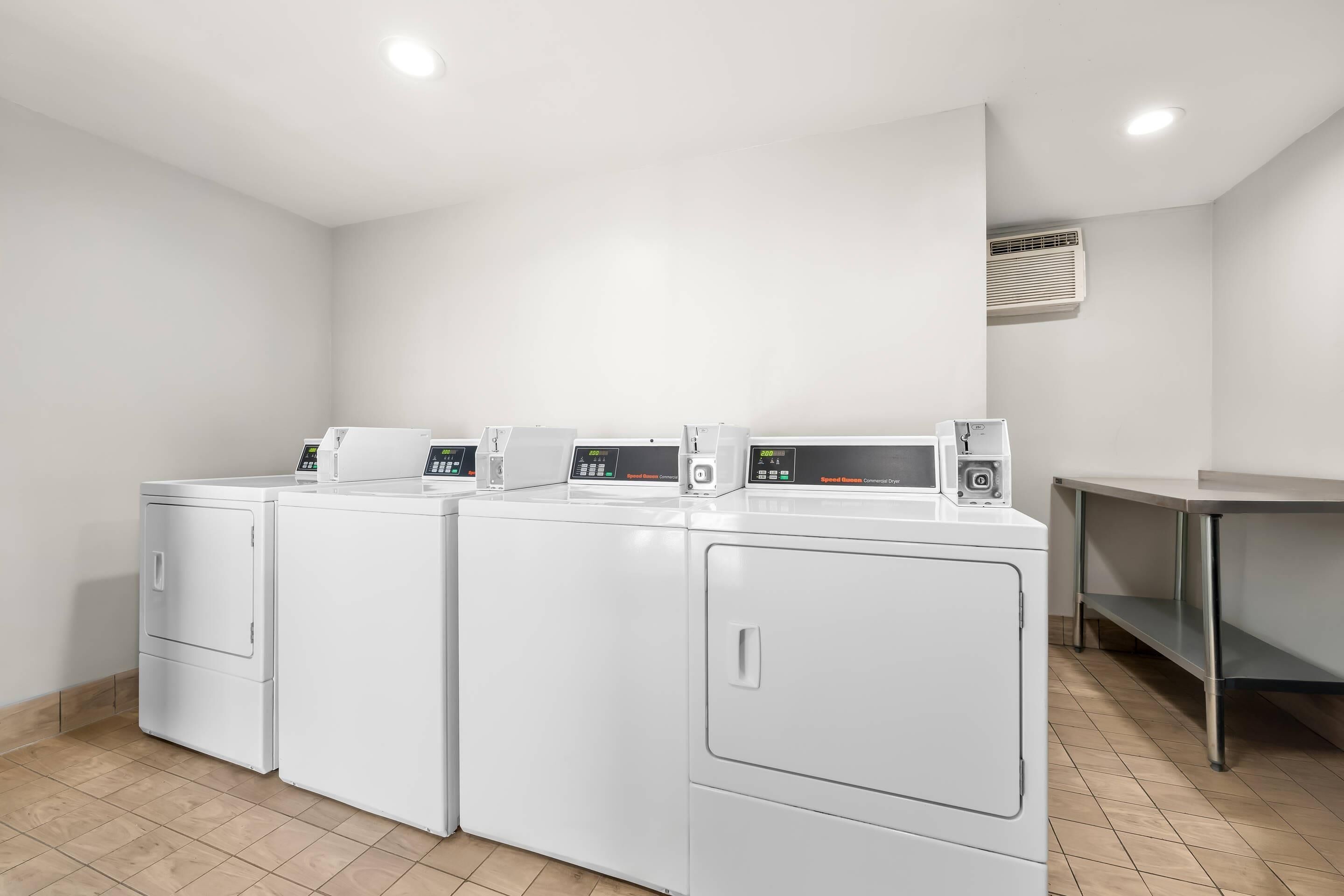 A laundry room with four white front-loading washers and dryers, a stainless steel table, and a vent on the wall.