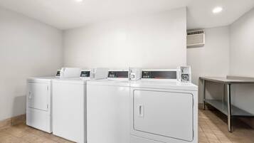 A laundry room with four white front-loading washers and dryers, a stainless steel table, and a vent on the wall.