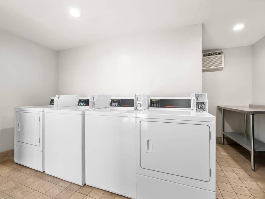 A laundry room with four white front-loading washers and dryers, a stainless steel table, and a vent on the wall.