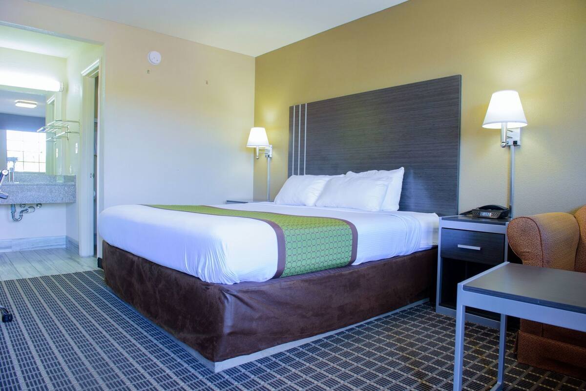 A hotel room with a bed, bedside lamps, a desk, a chair, and a bathroom visible in the background.
