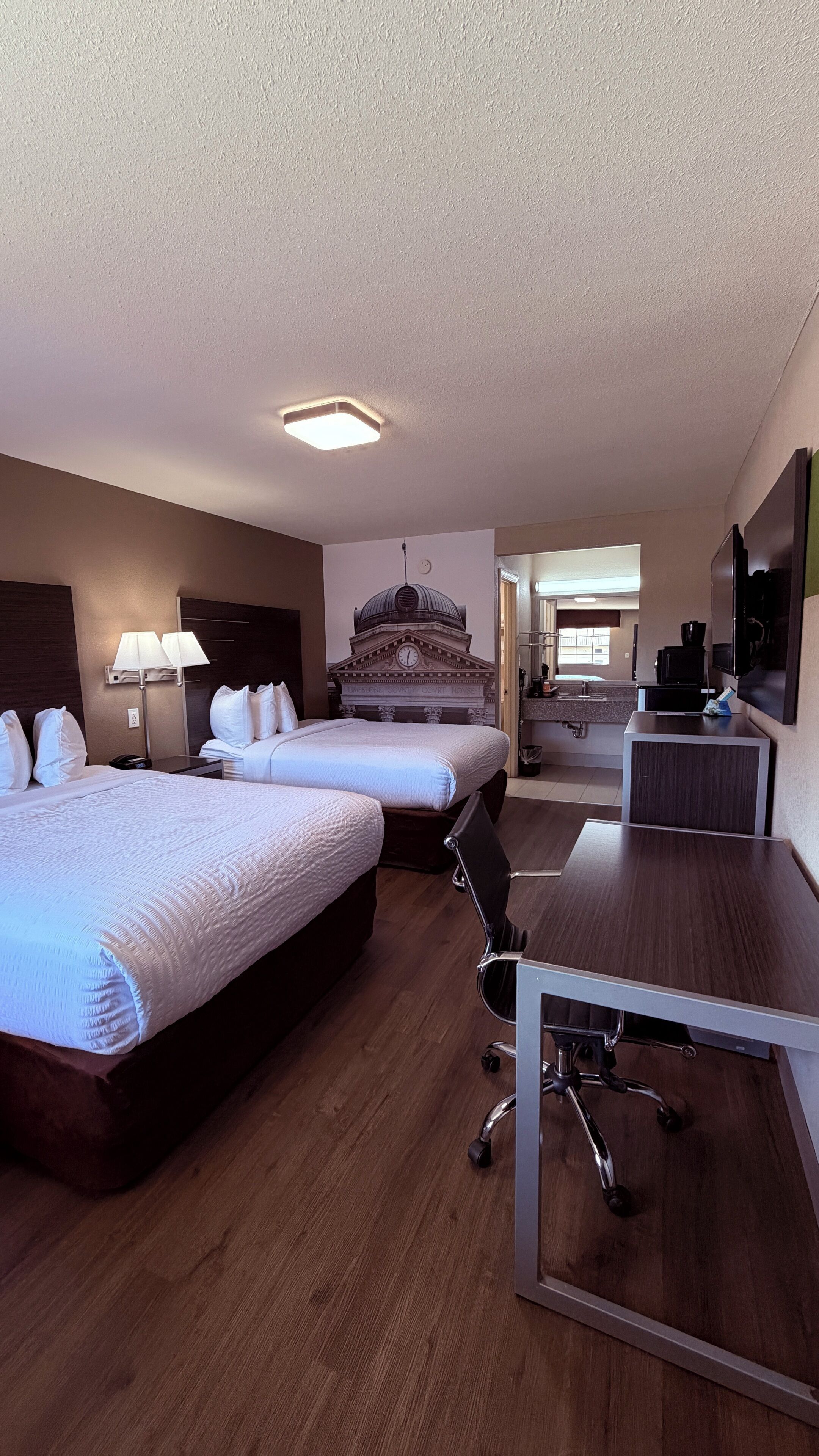 A hotel room with two beds, a desk, a chair, a lamp, a television, and a bathroom visible in the background.