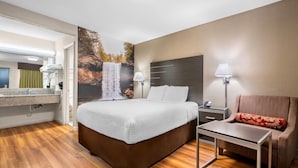 A hotel room with a bed, bedside table, chair, and a bathroom with a sink and mirror. - Clarion Pointe Athens I-65 (Athens)