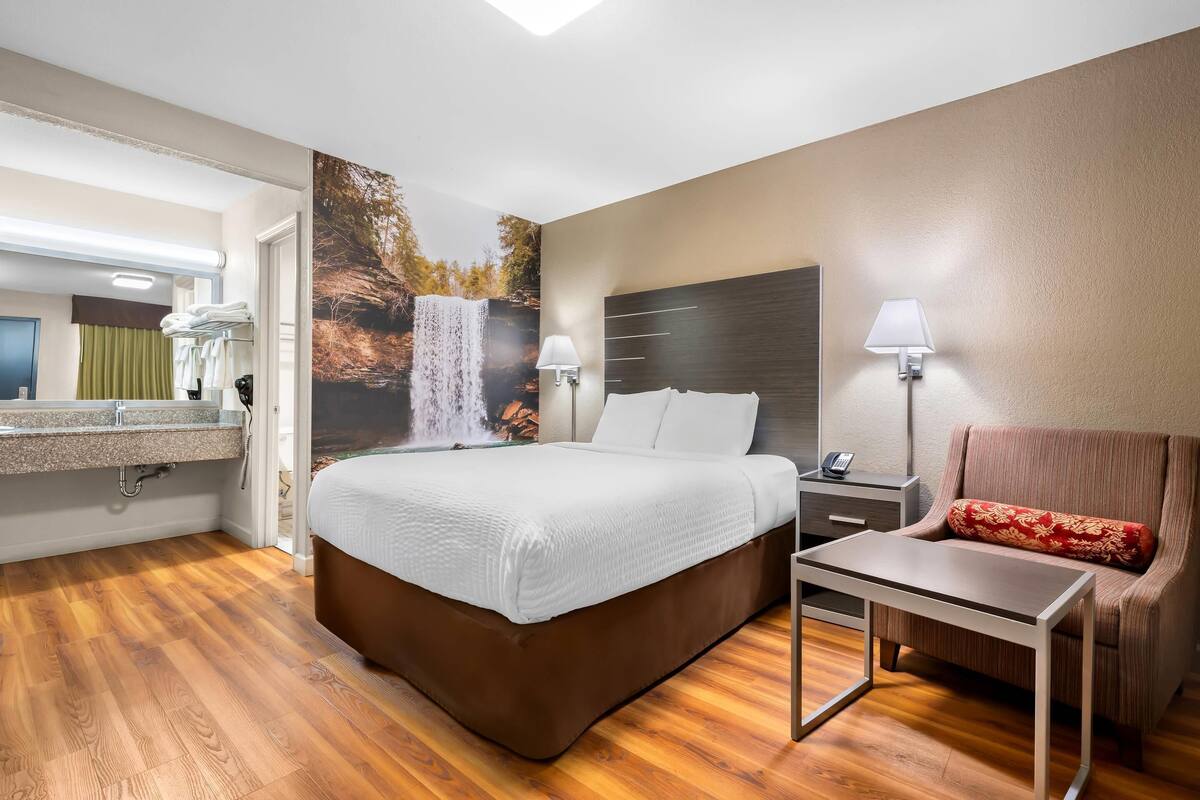 A hotel room with a bed, bedside table, chair, and a bathroom with a sink and mirror.