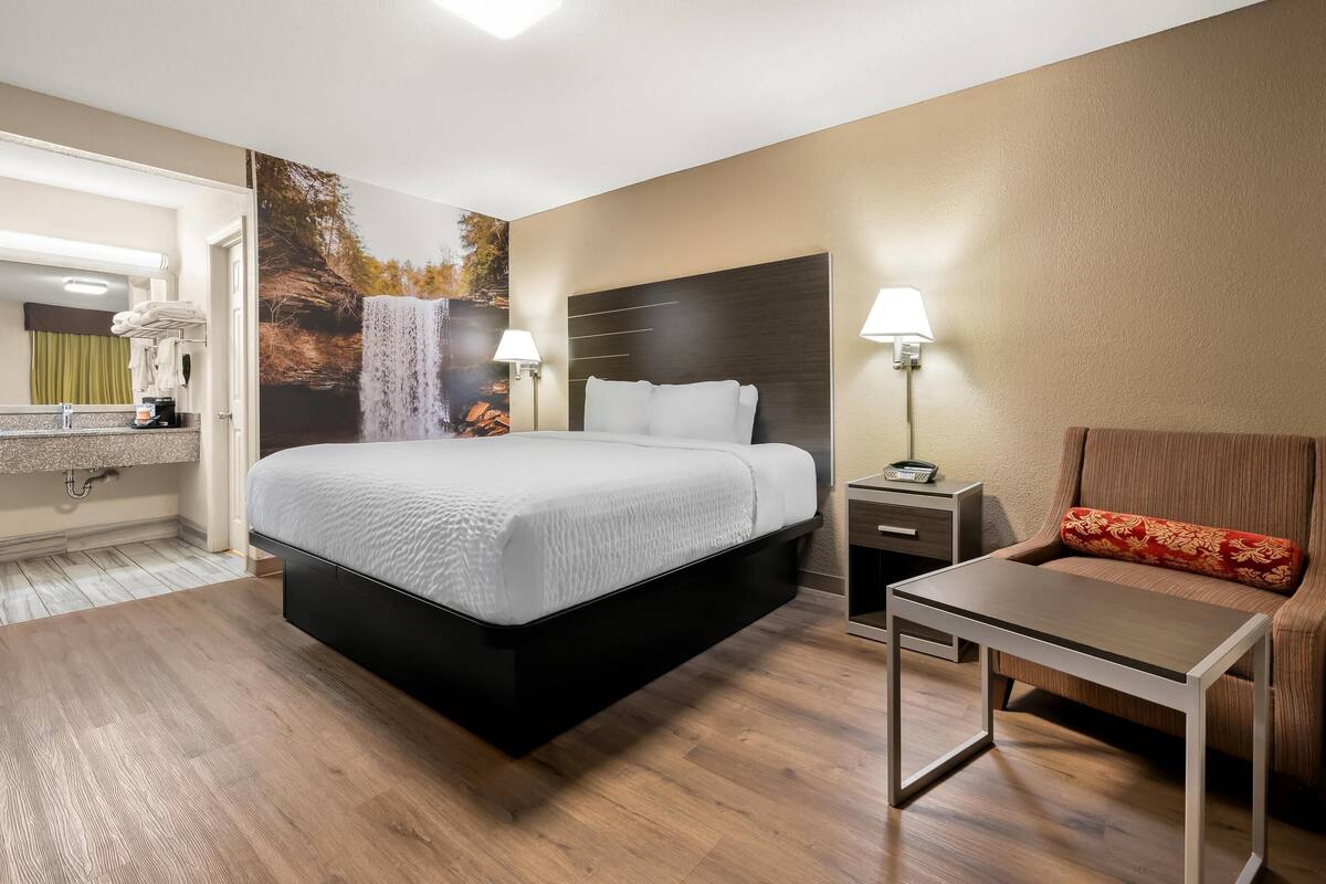 A modern hotel room with a bed, bedside table, chair, and a bathroom with a mirror and towel rack.