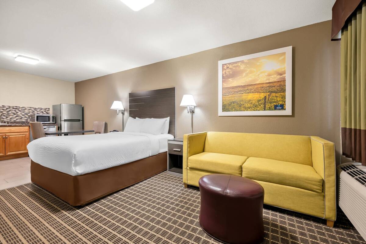 A hotel room with a bed, a sofa, a small table, a lamp, a picture on the wall, and a kitchenette.