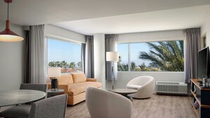 A modern hotel room with a large window, a sofa, a round dining table, and a TV.