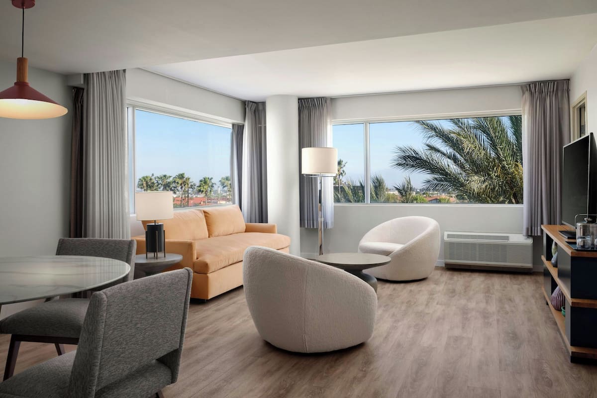 A modern hotel room with a large window, a sofa, a round dining table, and a TV.