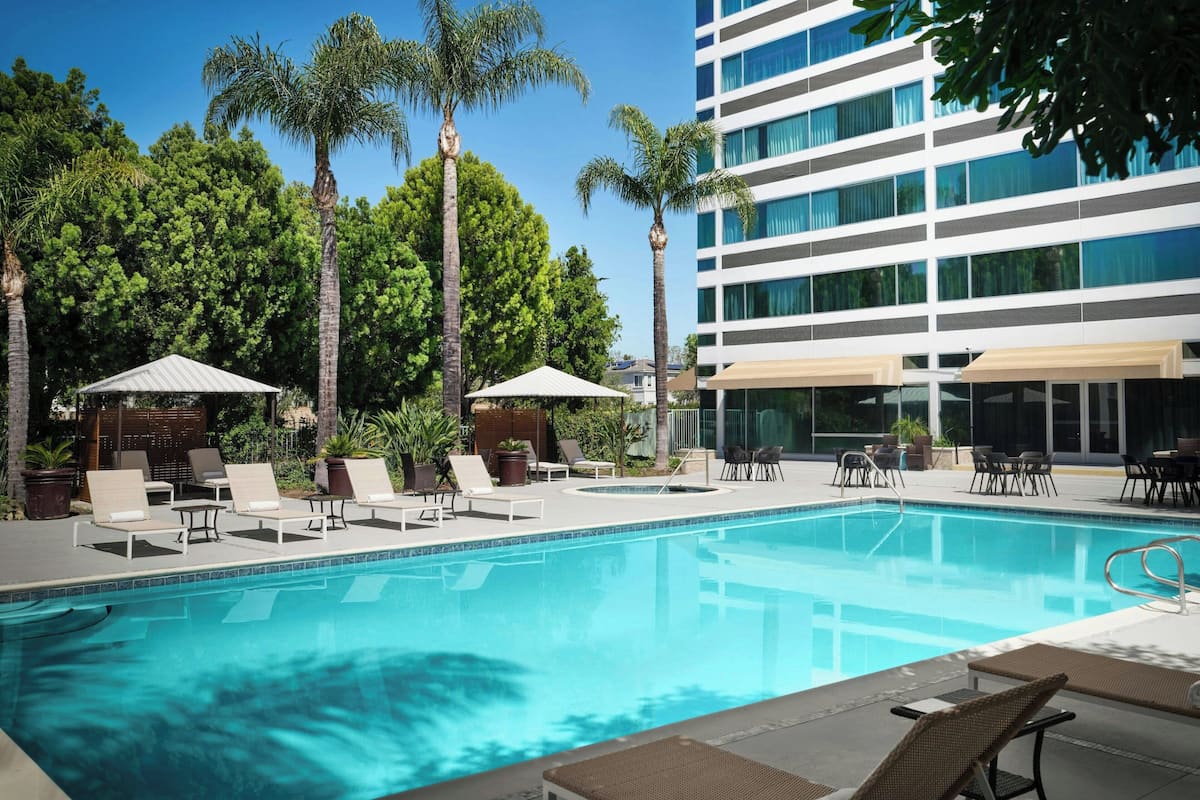A swimming pool with lounge chairs and umbrellas, surrounded by palm trees and a modern building.