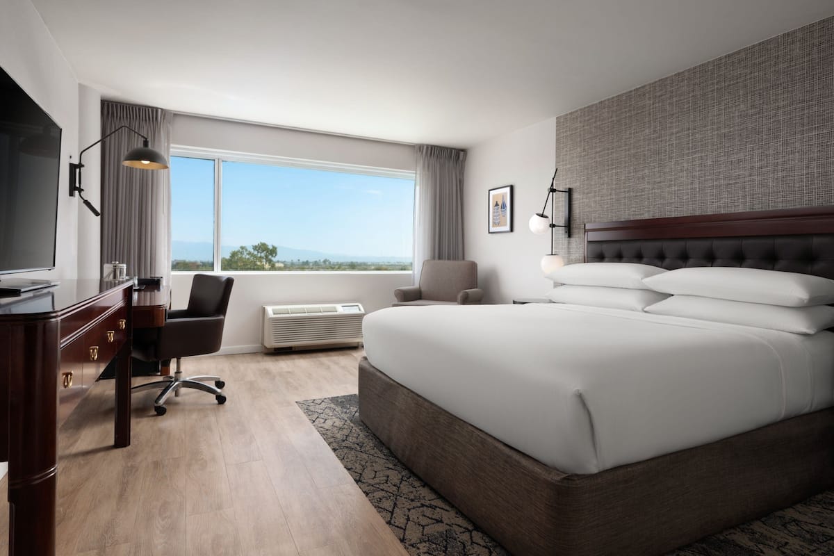 A modern hotel room with a large bed, a desk with a flat-screen TV, a chair, a large window with a view, and a wall-mounted lamp.