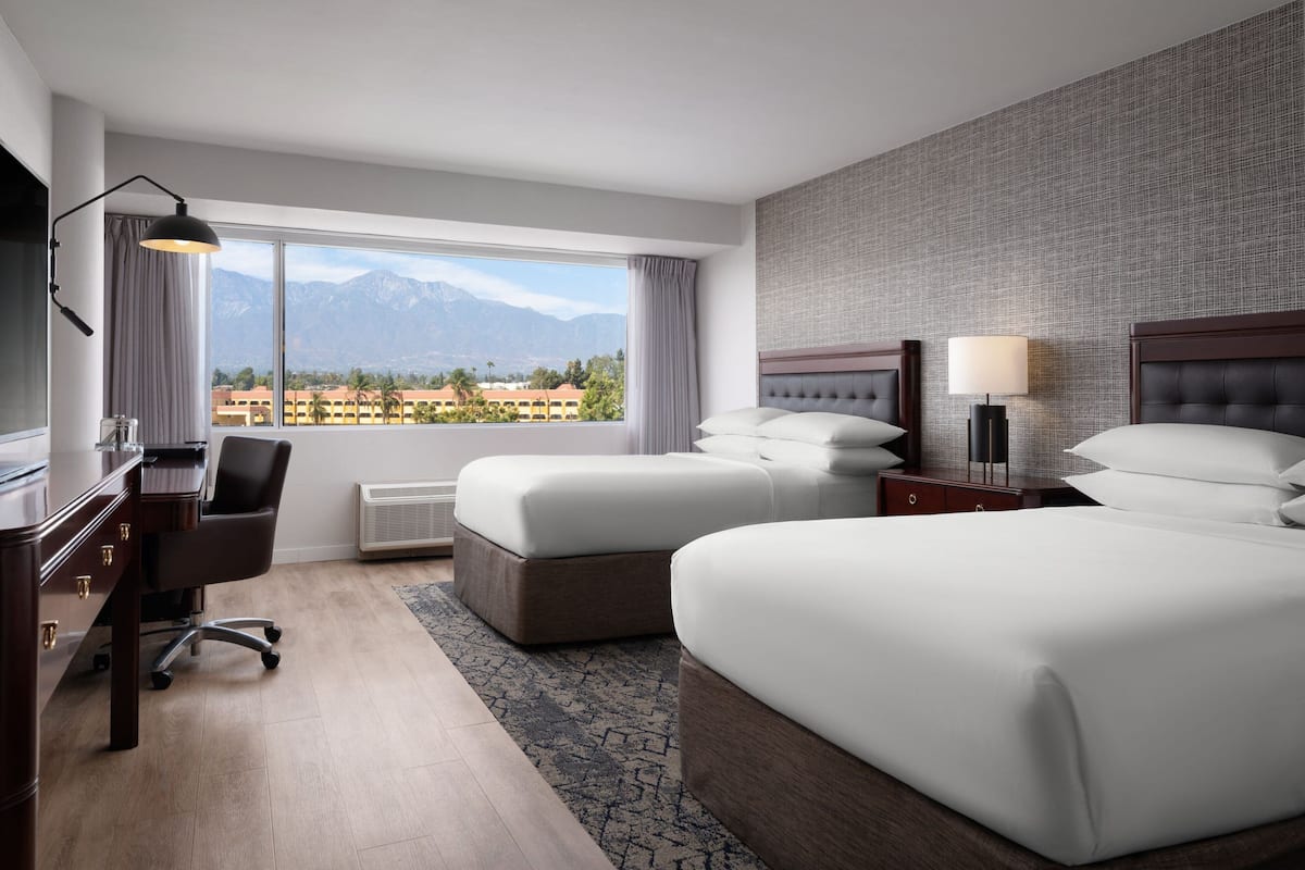 A hotel room with two beds, a desk, a chair, a lamp, and a view of mountains.