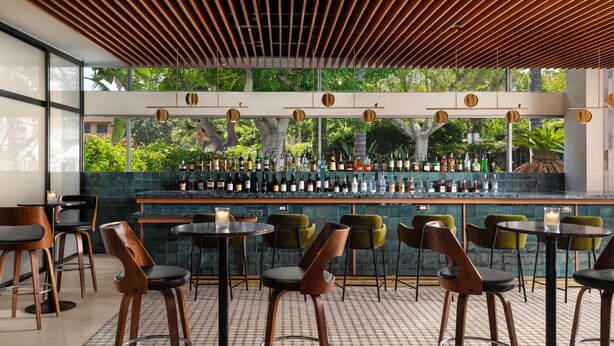 A modern bar area with wooden ceiling slats, green chairs, and a bar stocked with various bottles.