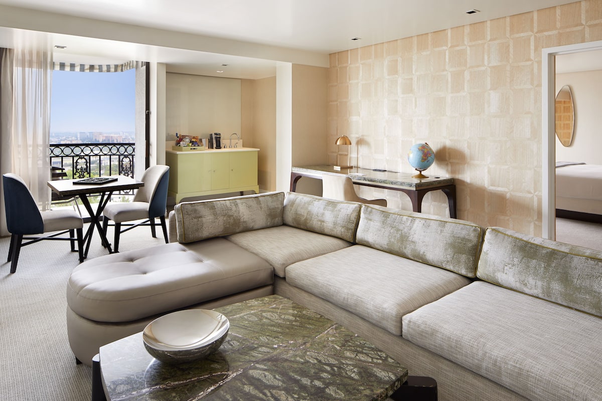A modern hotel room with a large sectional sofa, a dining table with chairs, a small kitchenette, and a balcony with a view.