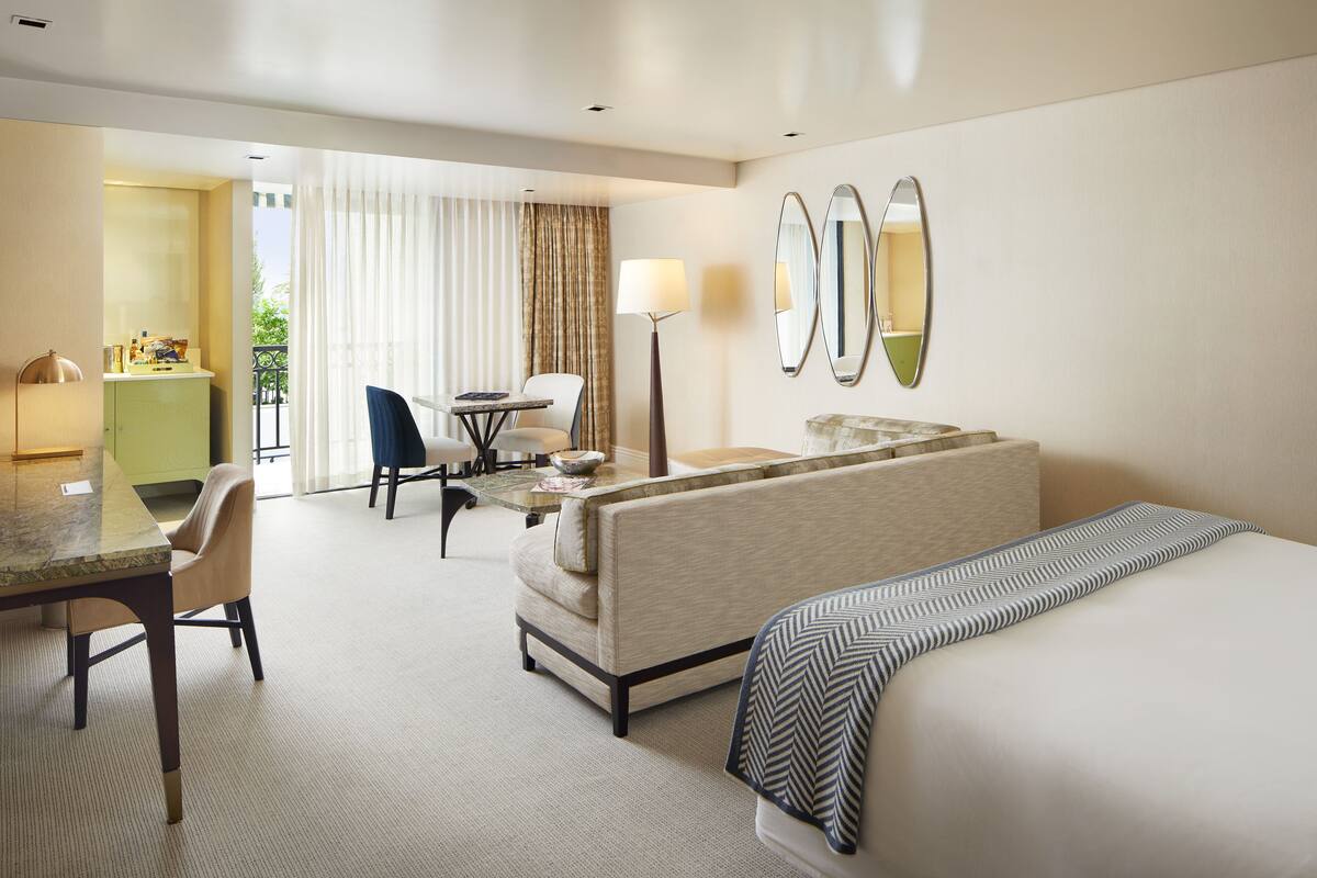 A hotel room with a bed, a sofa, a dining table with chairs, a lamp, and a view of the outdoors.