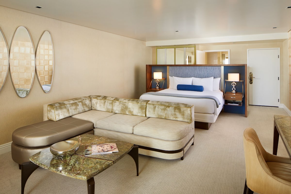 A hotel room with a sofa, a bed with a headboard, a nightstand, a table, and a chair.