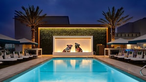 Una piscina con sillones de descanso y sombrillas, una pared con una obra de arte iluminada y palmeras.