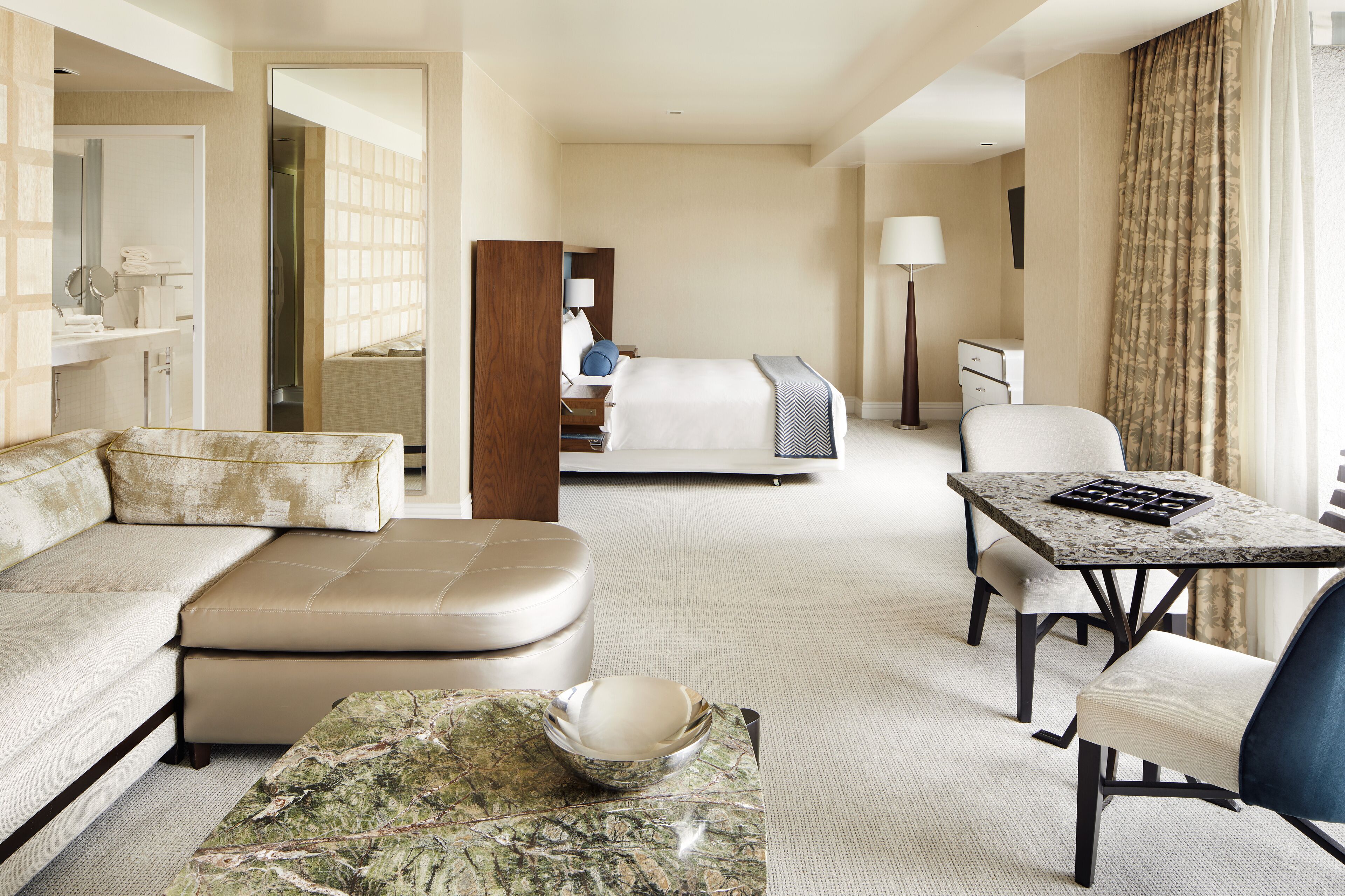 A hotel room with a sofa, a dining table, a bed, a mirror, a lamp, and a view of the outside.