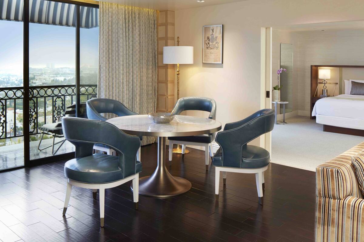 A hotel room with a balcony, a round dining table with chairs, a bed, and a small side table with a lamp.