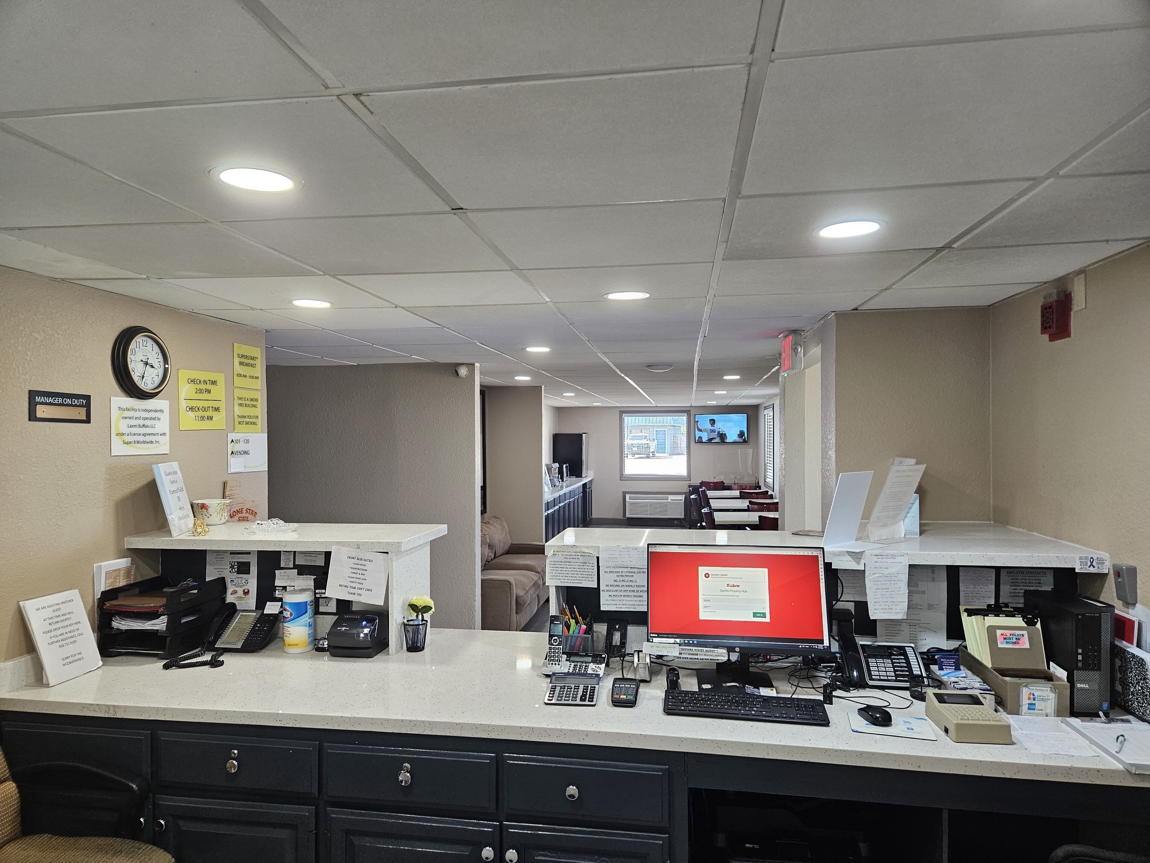 A hotel reception area with a desk, computer, and various office equipment. There are signs on the wall and a clock.