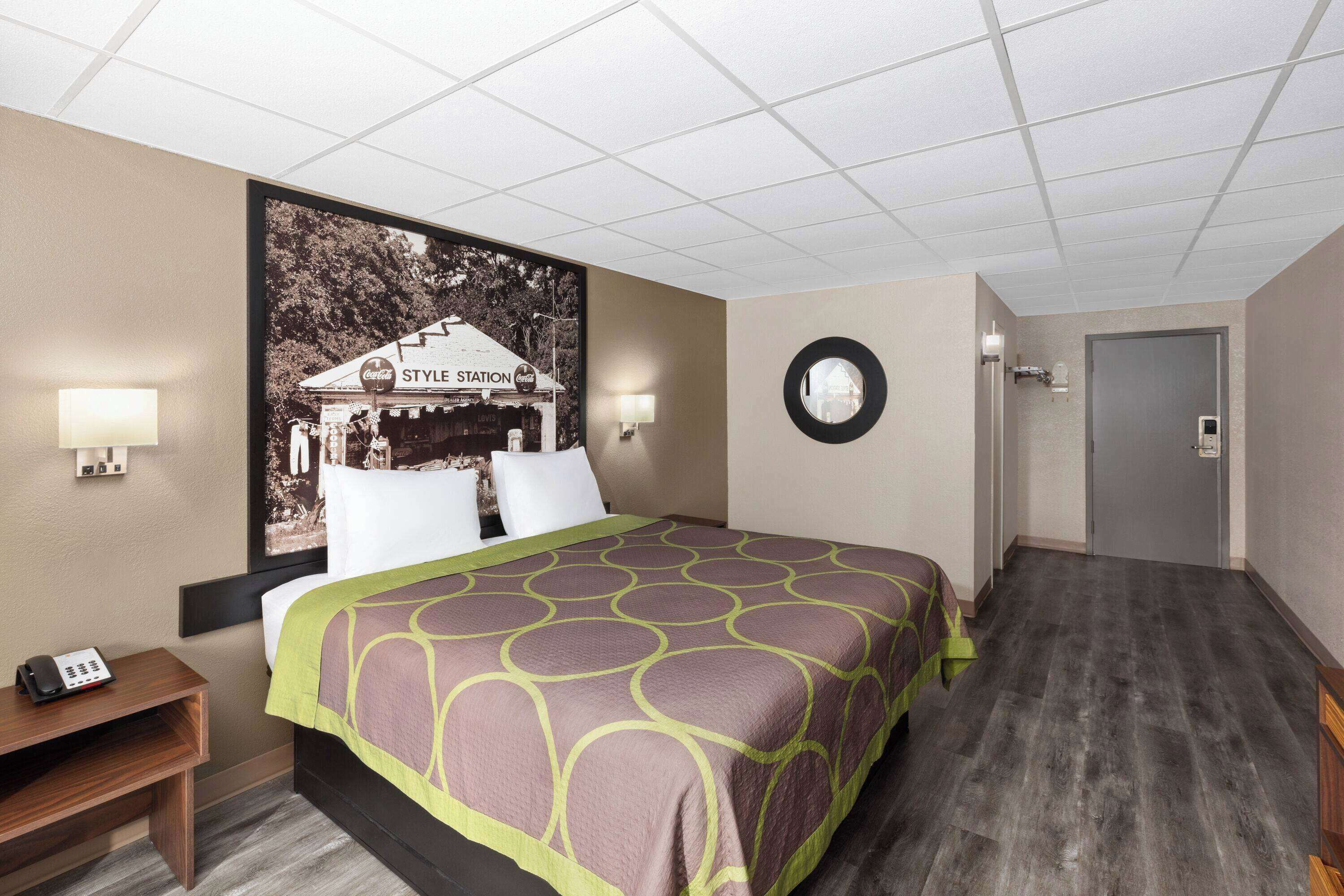 Habitación de hotel con una cama grande, una mesita de noche, una lámpara de pared y una puerta que da a otra habitación.